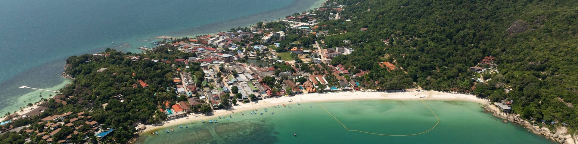 An aerial view of a coastal town with a beach, clear turquoise waters, and a mountainous backdrop.