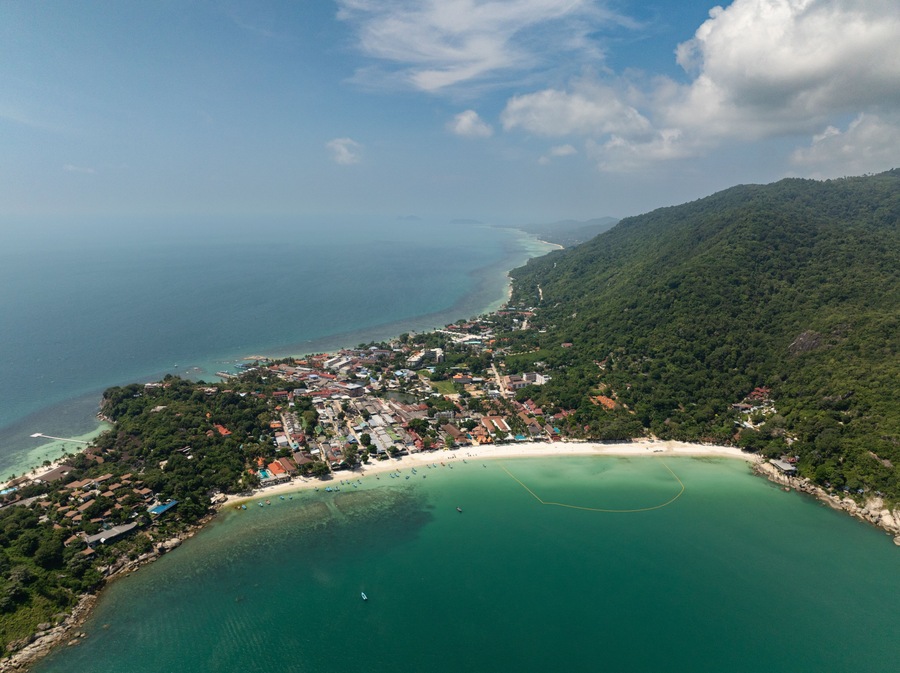 Lush green hills meeting the turquoise sea along a tropical coastline under a bright blue sky. Ko Pha Ngan, Thailand.