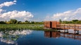 Un moll de fusta que s'endinsa en un estany ple de nenúfars, envoltat de vegetació exuberant i un cel blau clar.