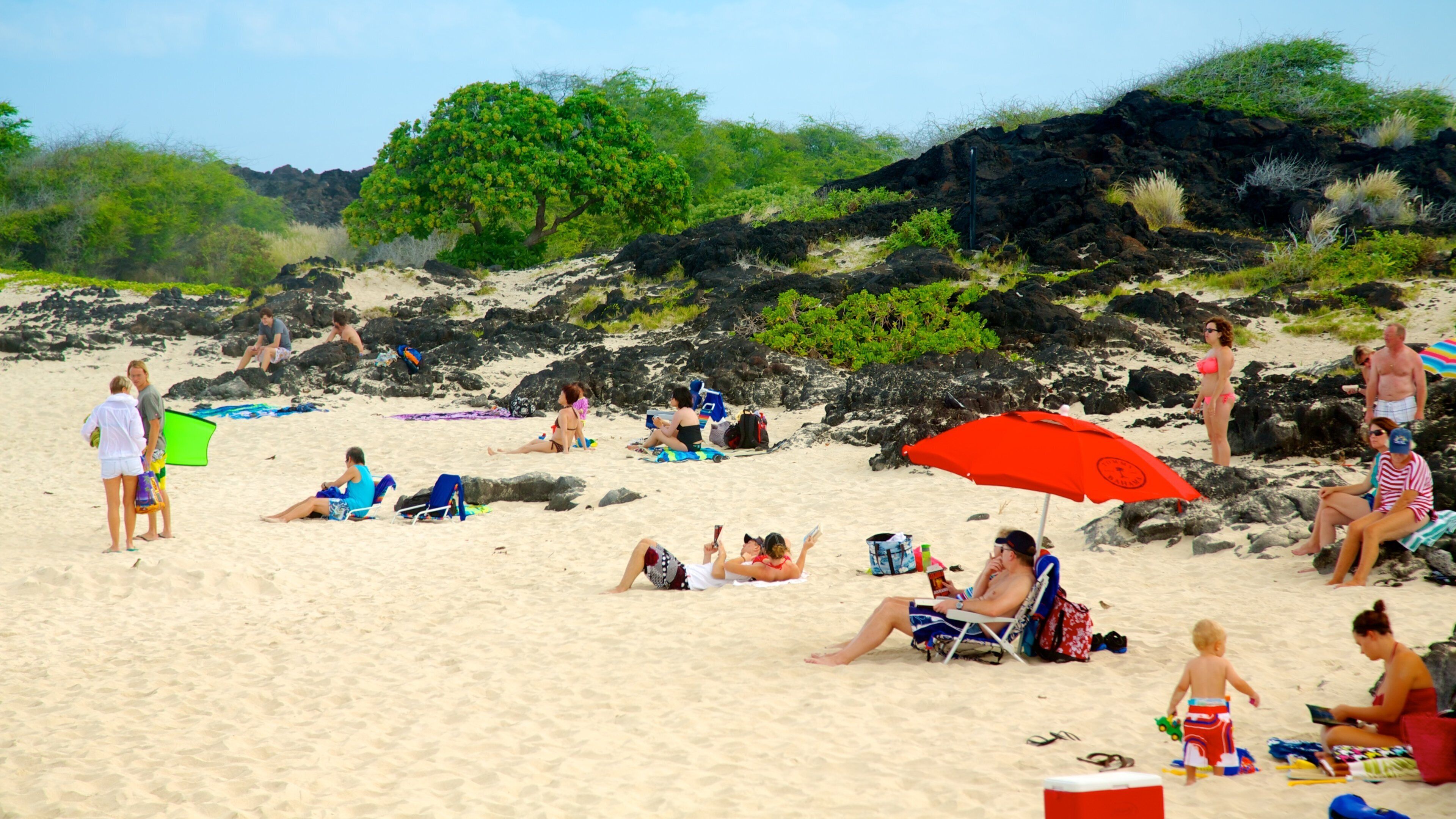 A sandy beach with people relaxing, a red umbrella, and rocky outcrops.