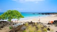 A beach with clear turquoise water, a large tree, and rocky outcrops.