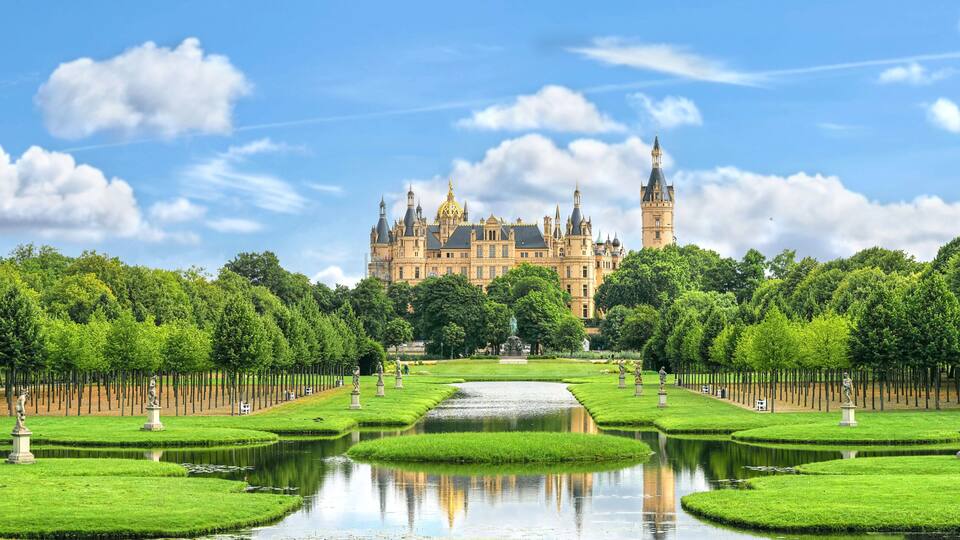 A grand castle with a prominent tower, surrounded by a meticulously maintained garden with a central pond and statues.
