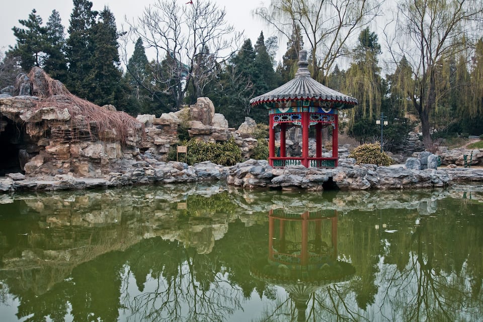 Un pavillon chinois traditionnel, situé au bord d’un étang, avec des formations rocheuses et des arbres en arrière-plan.
