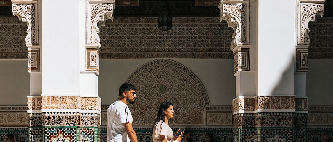 A man and woman walking past a building with intricate tile work and arches.