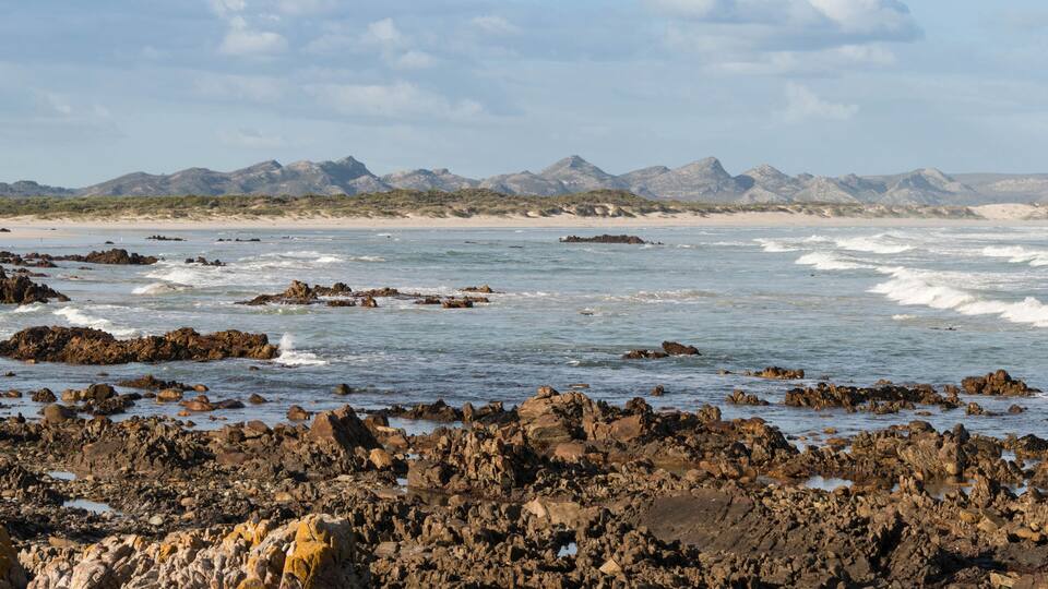 A rocky shoreline with waves crashing against the rocks and a distant mountain range under a partly cloudy sky.