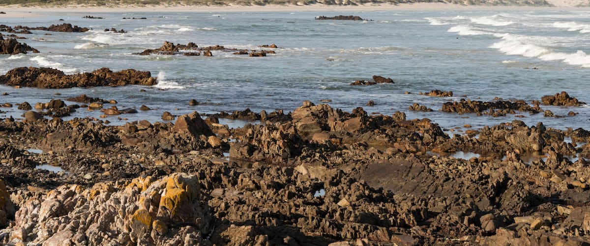 A rocky shoreline with waves crashing against the rocks and a distant mountain range under a partly cloudy sky.