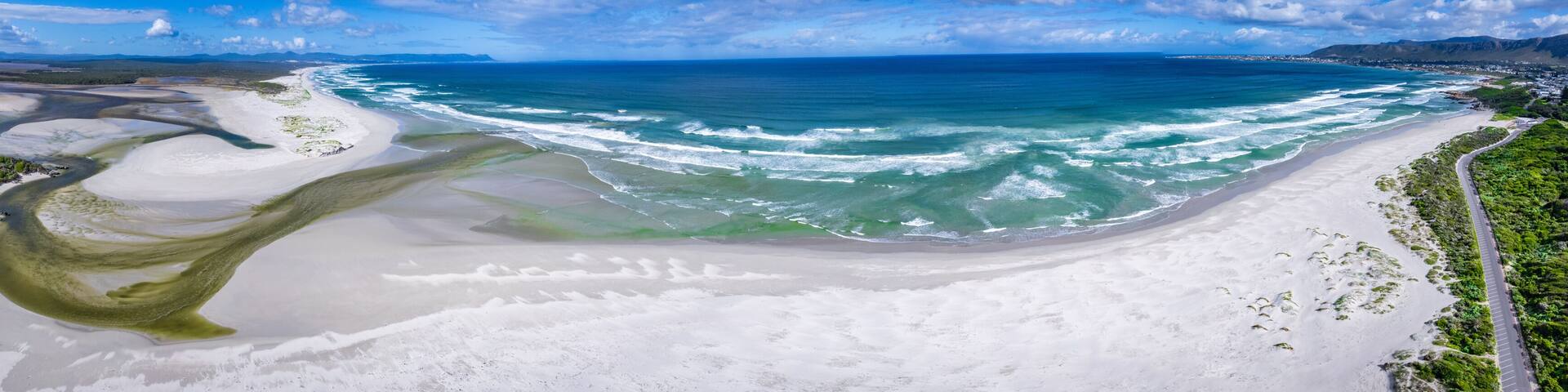 A coastal landscape with a sandy beach, clear blue ocean, and a road running parallel to the shore.