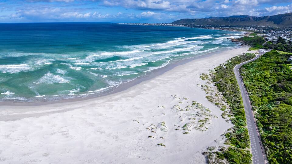 A coastal landscape with a sandy beach, clear blue ocean, and a road running parallel to the shore.