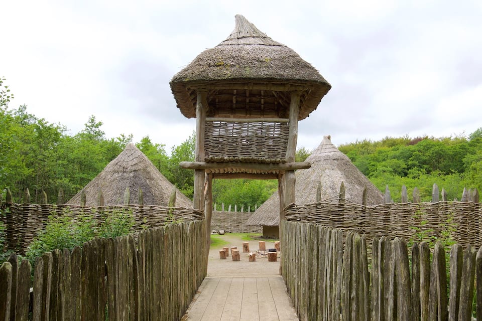 A traditional thatched-roof structure with a wooden walkway leading to it, surrounded by a wooden fence and lush greenery.