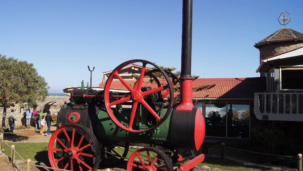 Ein alter Traktor mit roten Rädern und einem schwarzen Kasten, abgestellt in einer grünen Wiese, ein Gebäude im Hintergrund.
