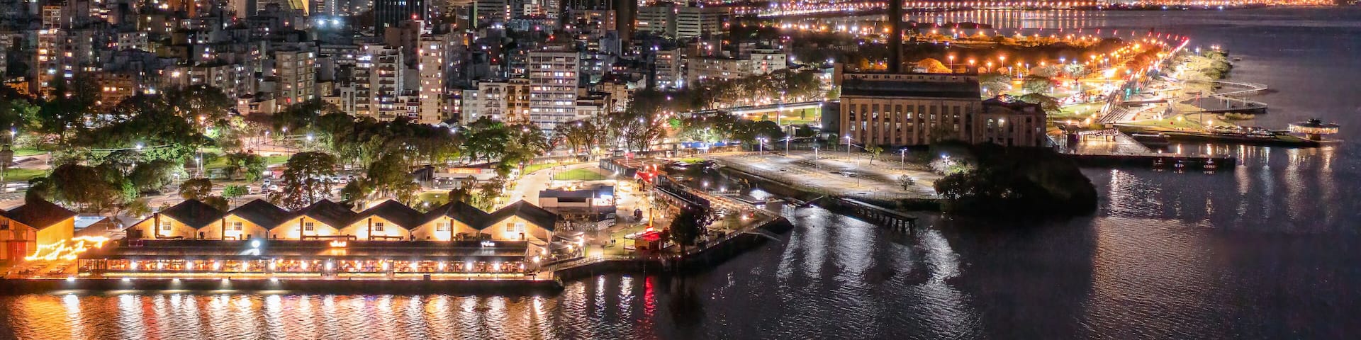 A nighttime cityscape with a waterfront promenade, illuminated buildings, and a prominent smokestack.