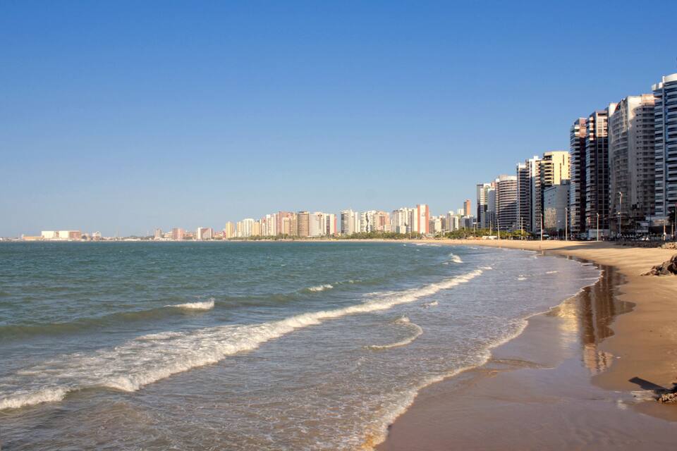 Una playa con olas, una orilla arenosa y un horizonte urbano de fondo.