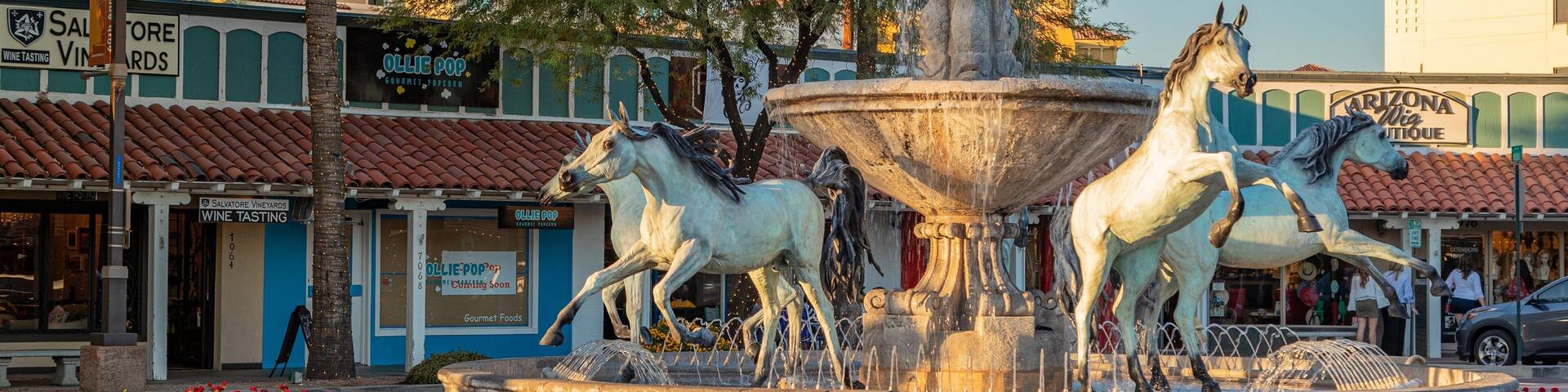 A fountain with three horses, surrounded by red flowers and storefronts.