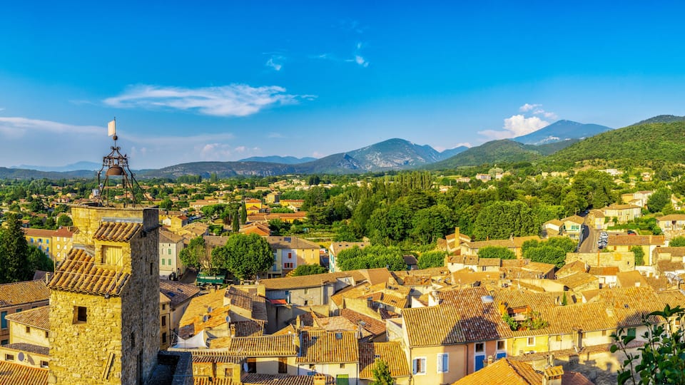 A historic town with terracotta-roofed buildings, a prominent bell tower, and a backdrop of green hills and mountains.