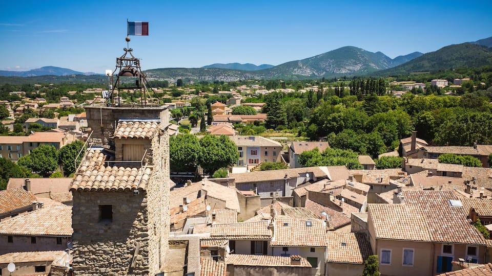 A town with stone buildings, a flag on a tower, and mountains in the background.