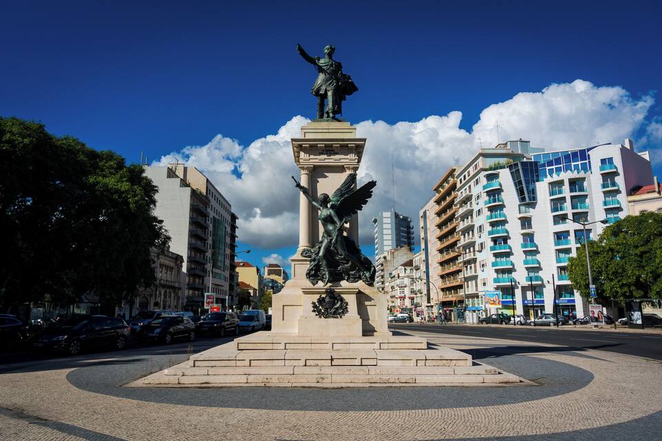 A statue of a figure with wings and a pedestal in an urban square with buildings and cars around.