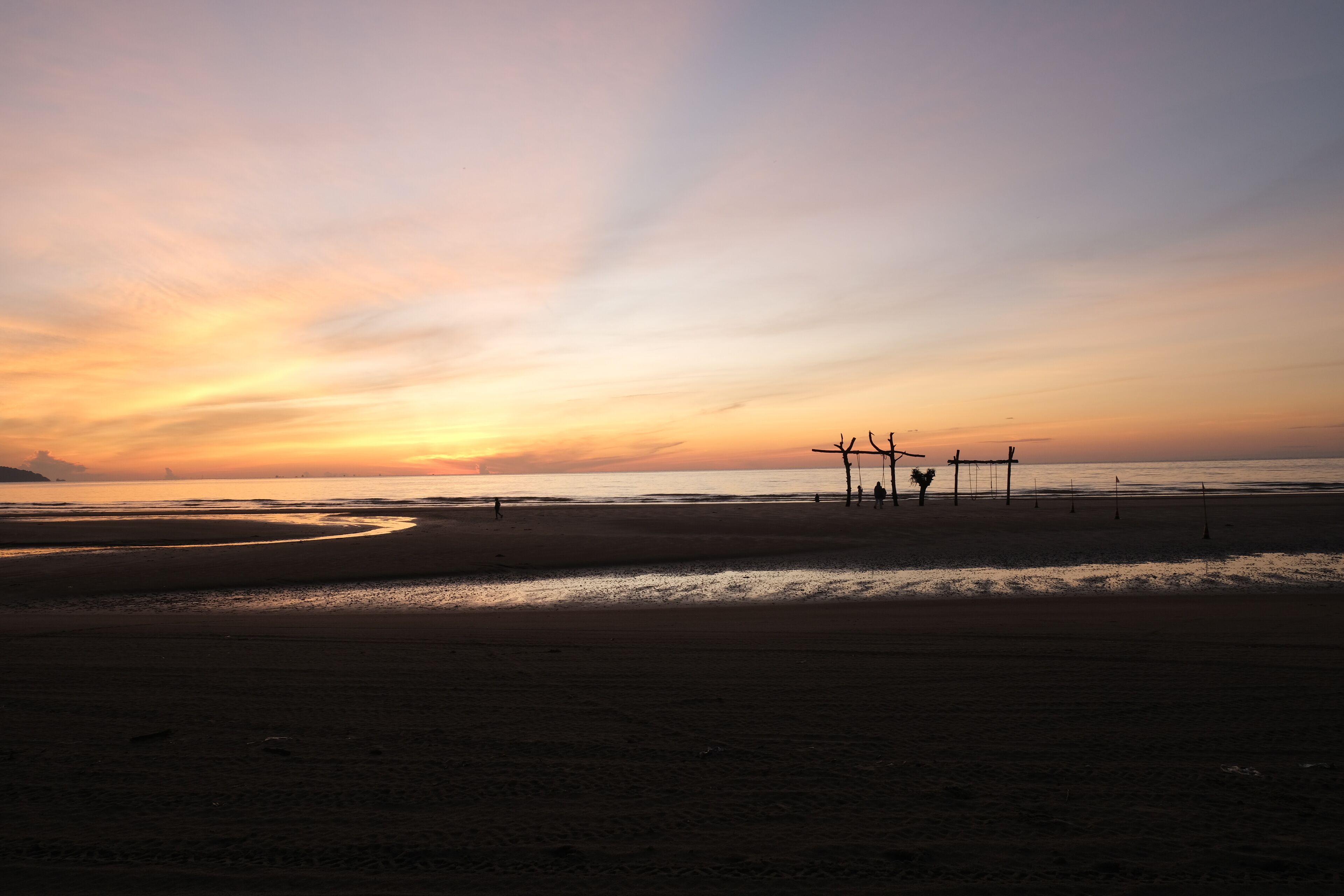 A beach at sunset with a calm sea, a few people, and a structure in the distance.
