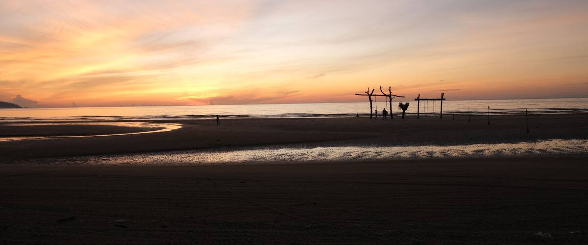 A beach at sunset with a calm sea, a few people, and a structure in the distance.