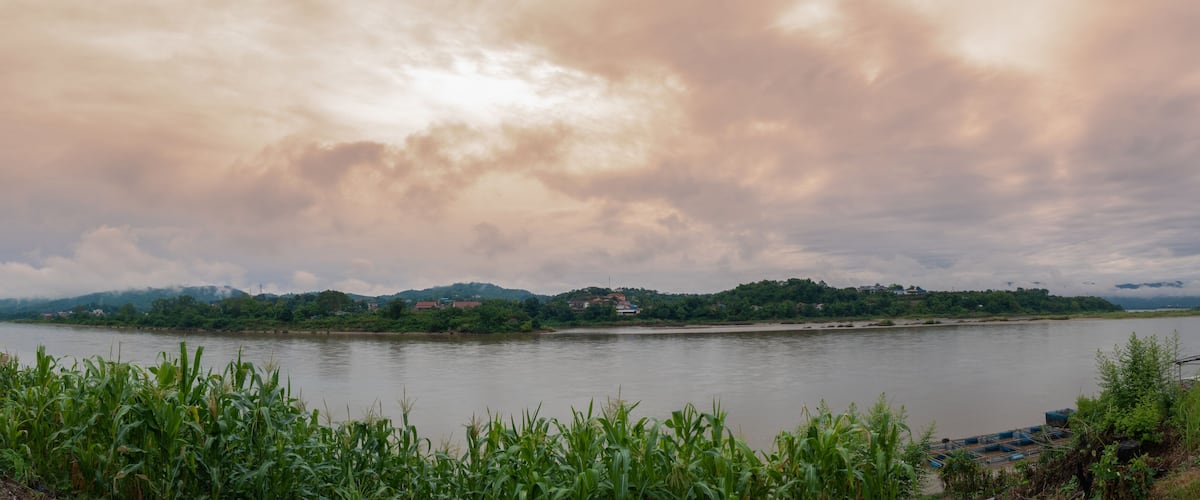 A river with a cloudy sky, green vegetation, and a distant hill with buildings.