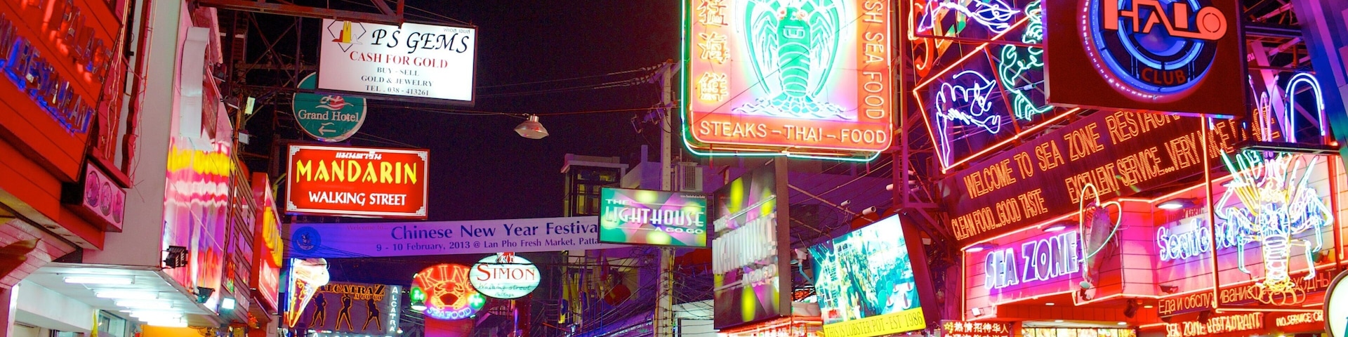Uma rua iluminada por néon com placas de restaurantes e lojas, incluindo 'Sea Zone' e 'Lobster Pot'.
