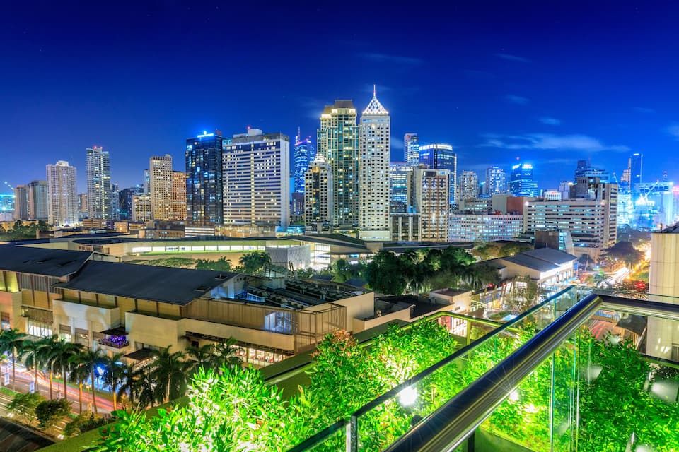Horizonte urbano noturno com edifícios iluminados e um jardim no terraço.