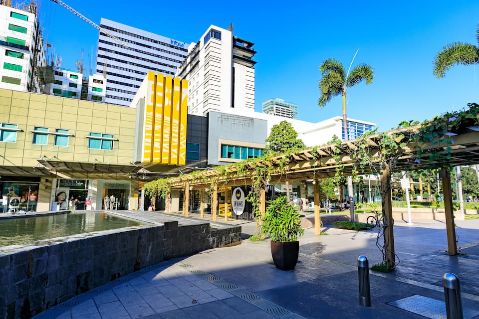 A city square with a water feature, modern buildings, and a covered walkway with greenery.