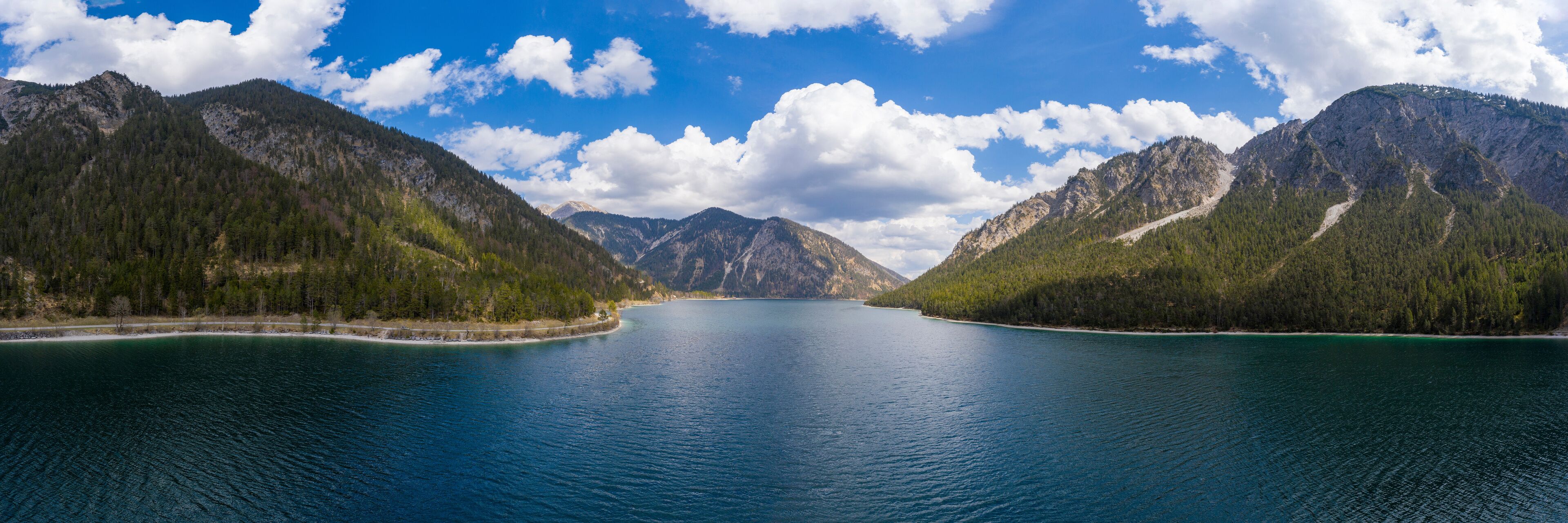 A calm lake surrounded by forested mountains under a partly cloudy sky.