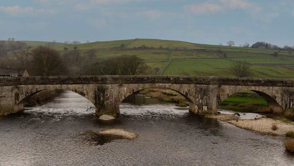 A stone bridge over a river with a rural landscape in the background.