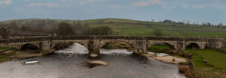 Un puente de piedra sobre un río, con un paisaje rural al fondo.