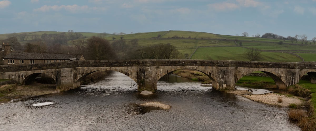 A stone bridge over a river with a rural landscape in the background.
