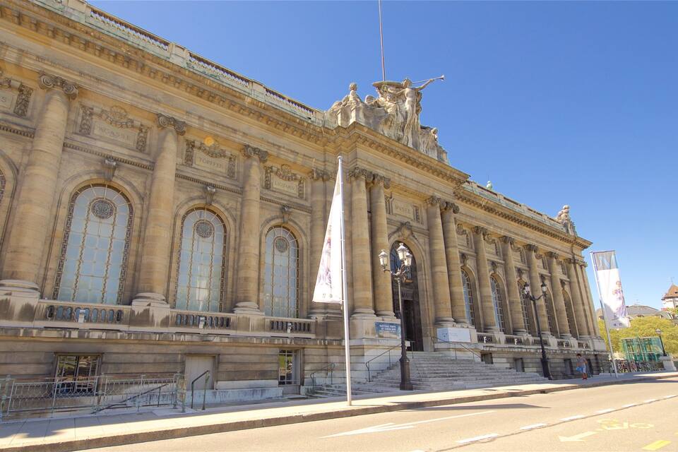 A grand neoclassical building with large arched windows and columns, a prominent statue, and a clear blue sky.