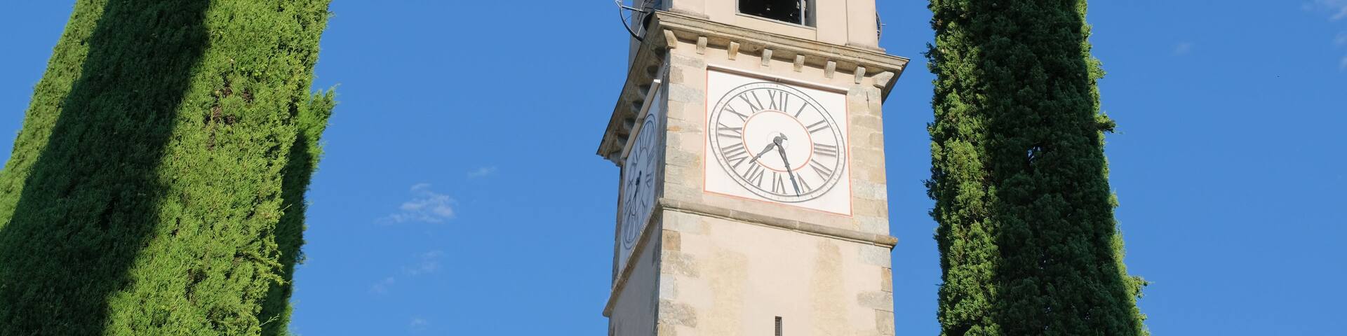 A clock tower with a dome and a bell, surrounded by tall, narrow trees against a clear blue sky.