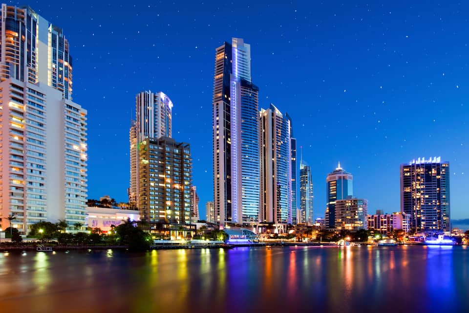 A city skyline at night with illuminated buildings and a calm river reflecting the lights.