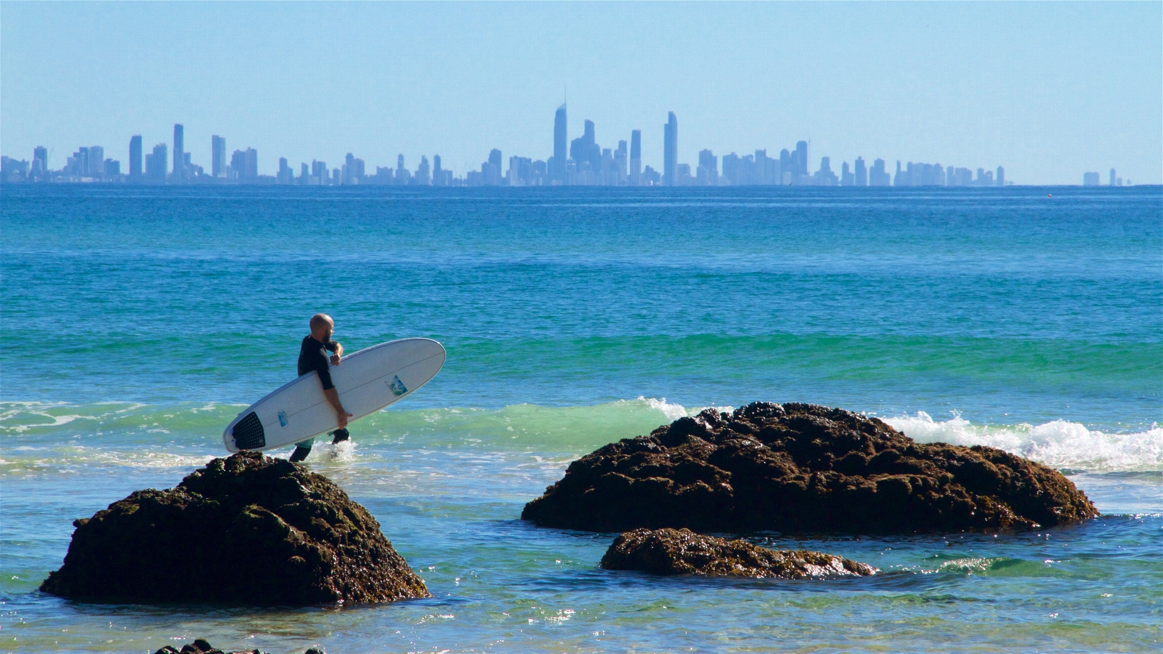 A surfer walking between rocks with a city skyline in the background.