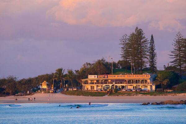 Un restaurant situé en bord de mer, avec une enseigne indiquant « Rainbow Bay SLSC » et une vue sur les surfeurs dans l’eau.