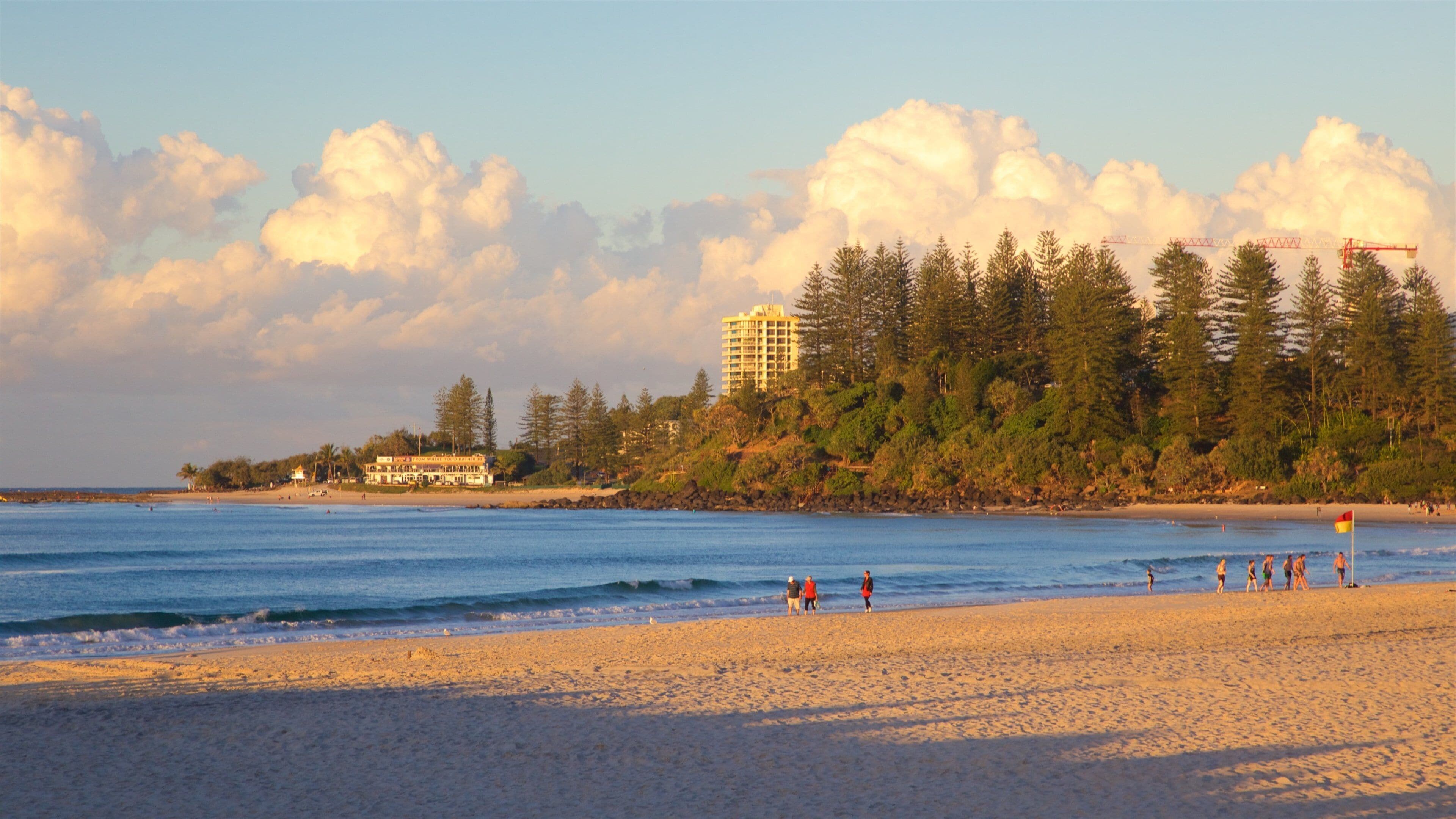 A beach with people walking, a building, and a forested hill.