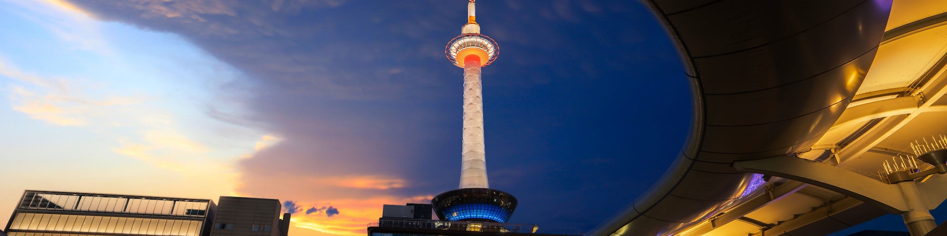 Paisagem urbana ao anoitecer, com uma torre imponente iluminada contra o céu do pôr do sol.