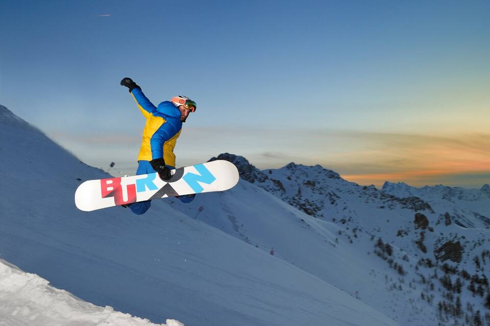 Un snowboarder en pleno vuelo con montañas al fondo durante el atardecer.