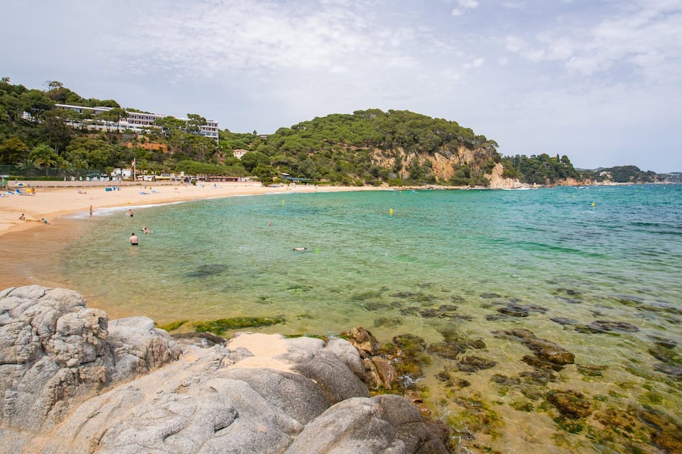 A beach with clear turquoise water, rocky shoreline, and buildings on a hillside.