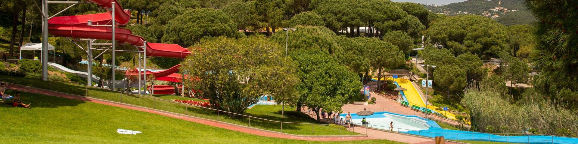 A park with a red water slide, a swimming pool, and people sunbathing on the grass.