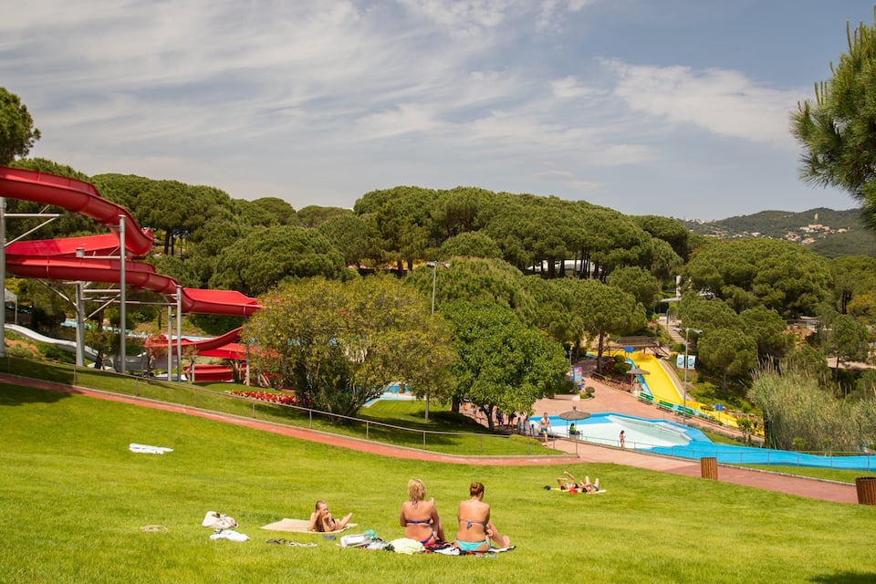 Um parque com um escorrega aquático vermelho, uma piscina e pessoas tomando sol na grama.