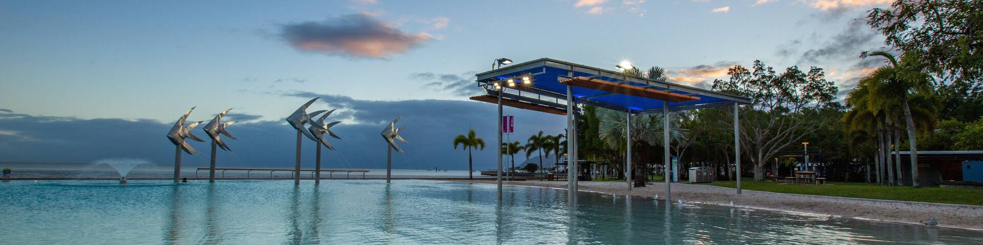 A swimming pool with wind sculptures, a covered pavilion, palm trees, and a sunset sky.
