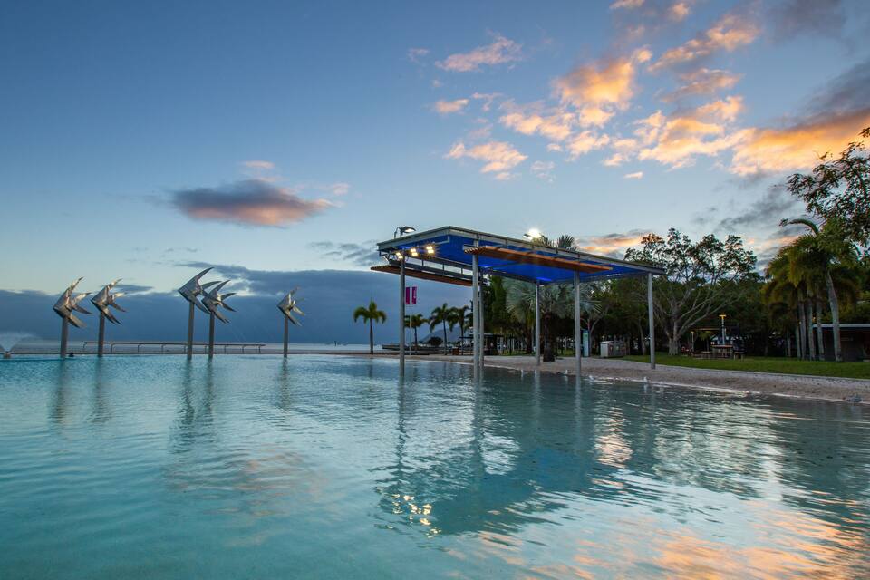 Una piscina con esculturas eólicas, un pabellón cubierto, palmeras y un cielo al atardecer.