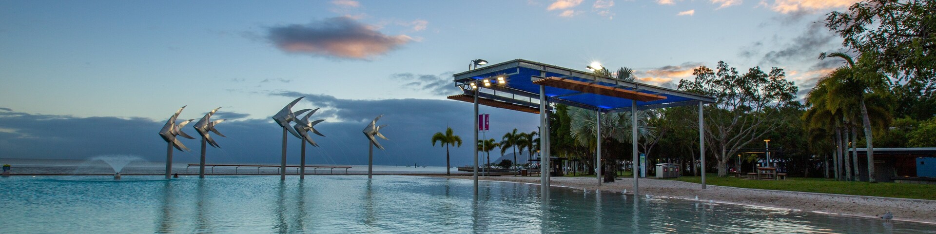 Piscina com esculturas de vento, um pavilhão coberto, palmeiras e um céu ao pôr do sol.