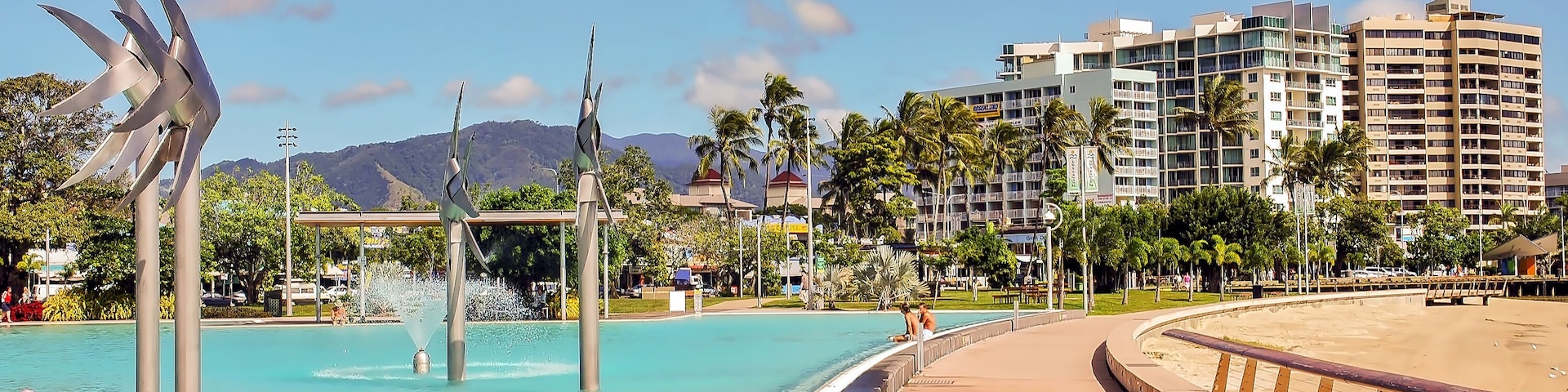 Un parque costero con piscina, palmeras y edificios modernos al fondo.