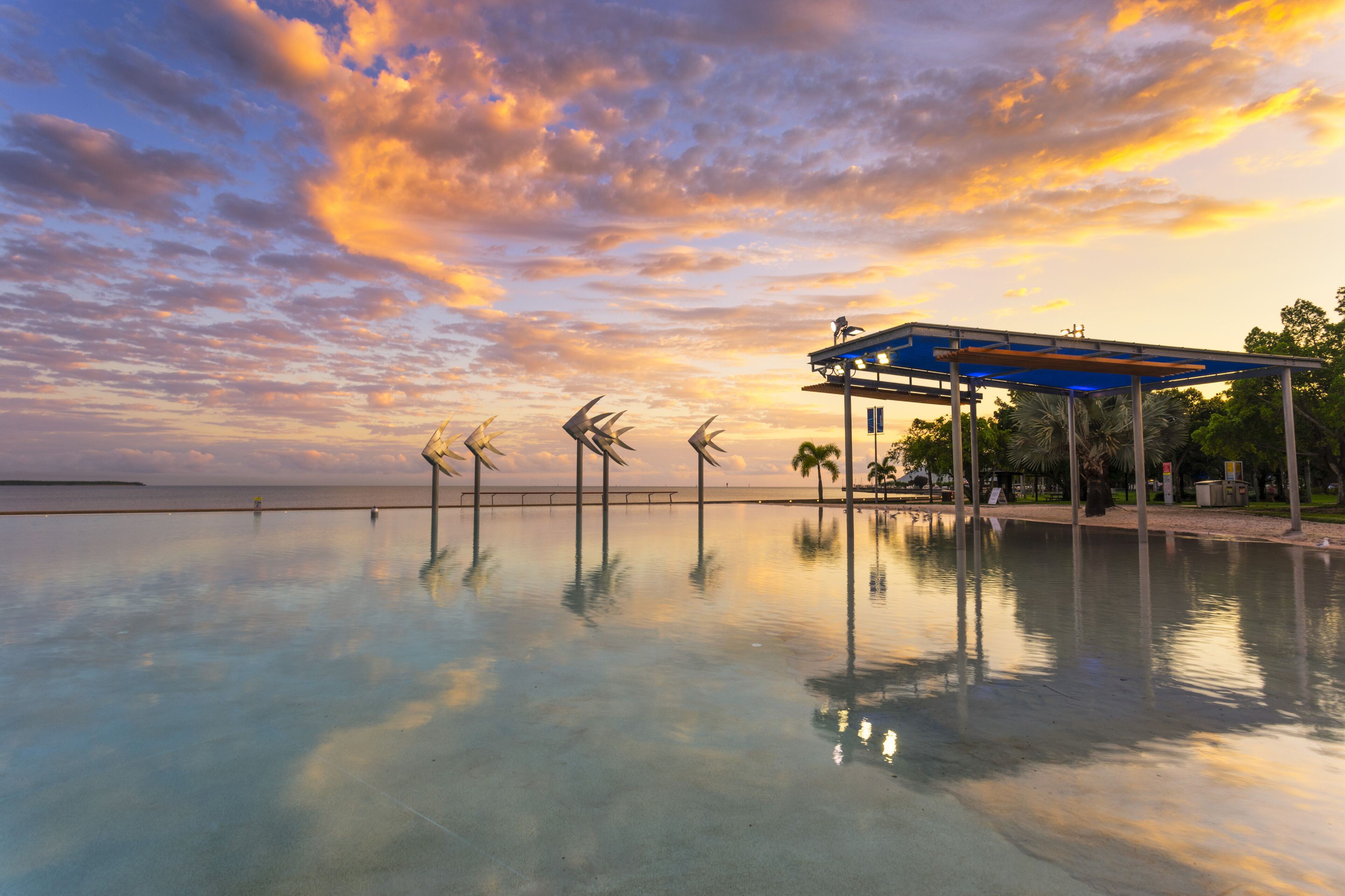 A waterfront with a reflective pool, a blue-roofed pavilion, and wind sculptures.