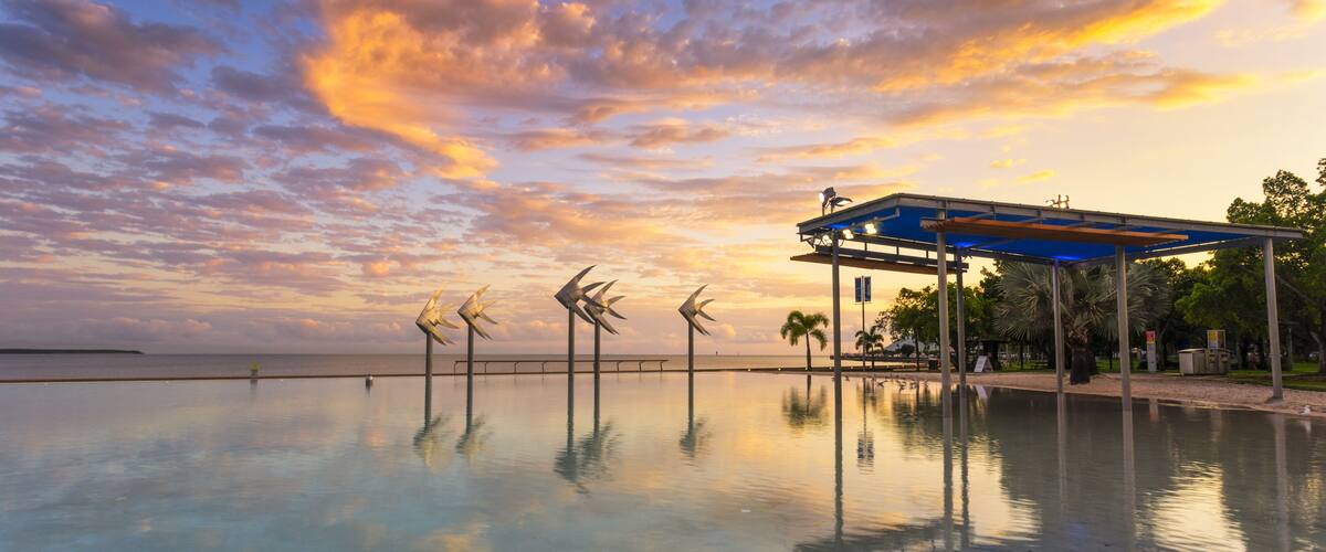 A waterfront with a reflective pool, a blue-roofed pavilion, and wind sculptures.