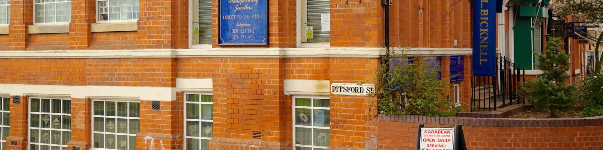 A brick building with signs for G.L. Bicknell & Sons Ltd, a jewellery store, and a sign for a restaurant serving jerk chicken and curry.