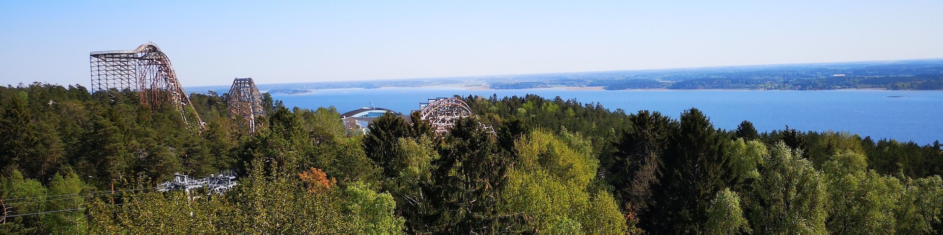 A coastal landscape with a roller coaster, dense forest, and a clear view of the ocean.