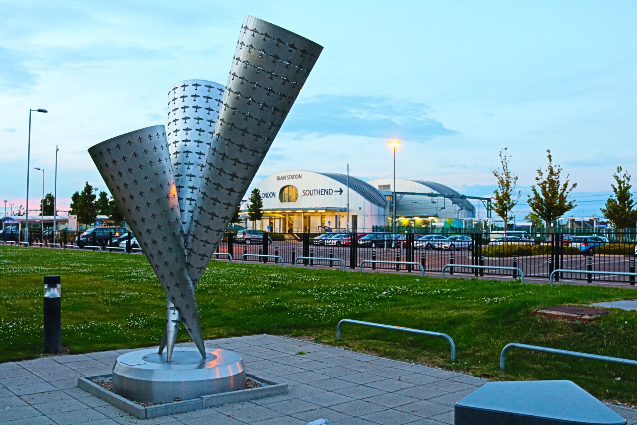 A modern sculpture in a public square, with a train station and cars in the background.